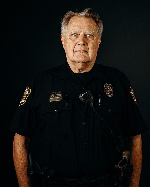 Police officer in dark uniform, standing with stern expression against a dark background.