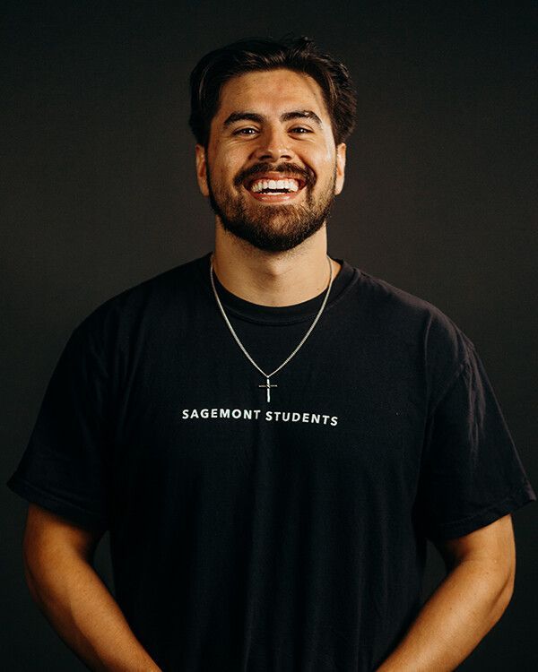 Man with a beard smiling, wearing a red polo shirt, against a black background.