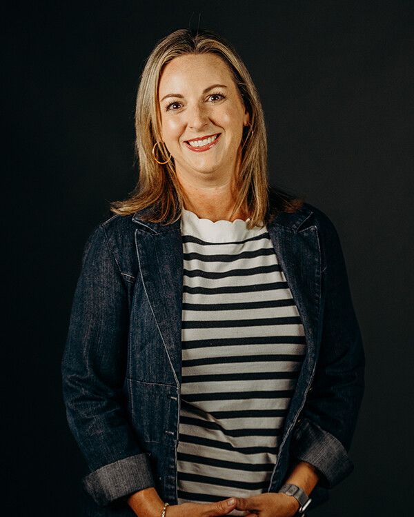 Woman smiling, wearing a striped shirt and denim jacket, posing against a dark background.