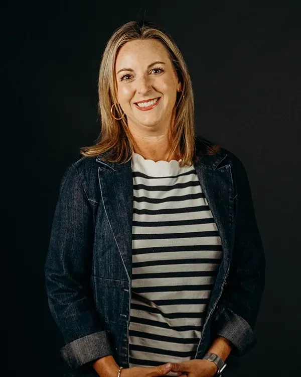 Woman with blonde hair wearing a denim jacket and striped shirt, smiling in front of a dark backdrop.