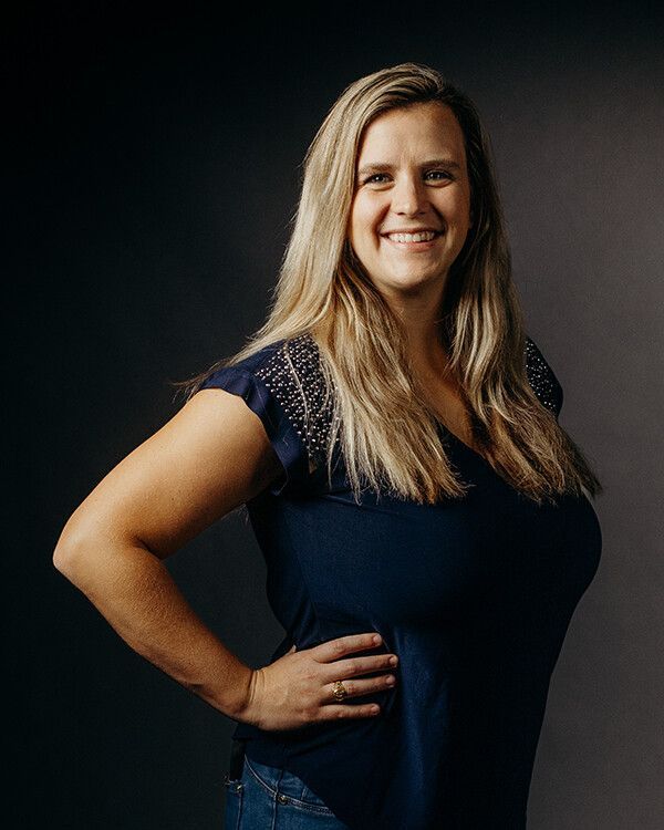 Woman with blonde hair, smiling, wearing a blue top, hand on hip, standing against a dark background.