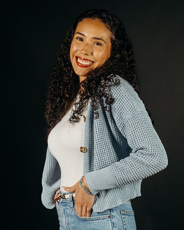 Woman smiling, wearing a white top and blue cardigan, posing with hand in pocket, against a black backdrop.