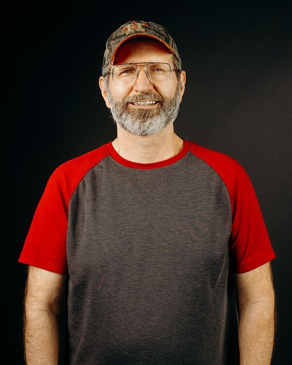 Man with glasses and a beard wearing a camouflage hat and a gray and red baseball shirt, smiling.