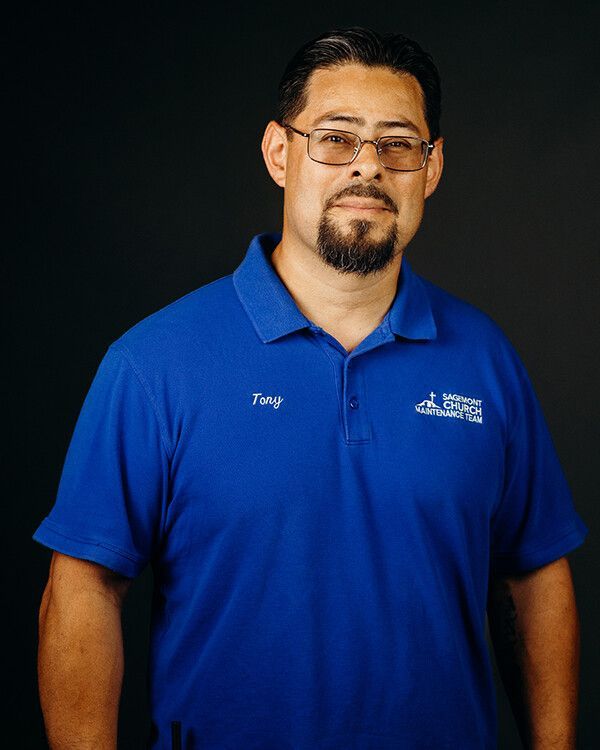 Man in blue polo shirt and glasses, standing against a dark background, smiling.