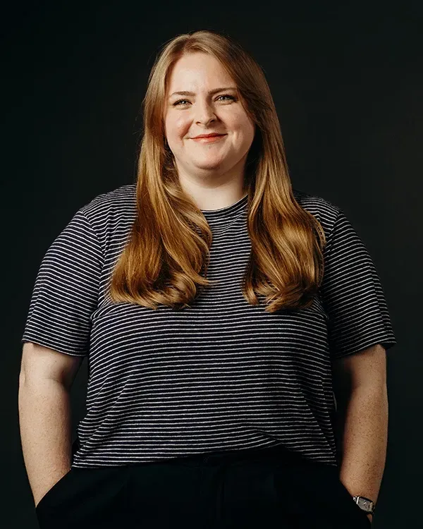 Woman with long blonde hair, smiling, wearing a striped shirt and dark pants, hands in pockets.