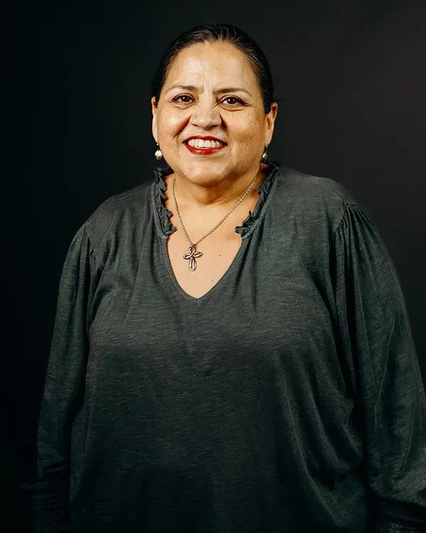 Woman smiling, wearing a gray top and a necklace, against a black background.