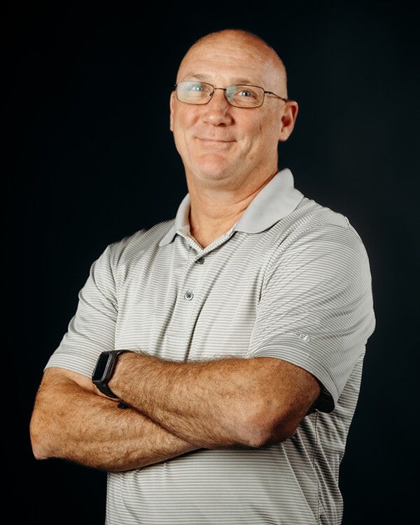 Bald man in glasses, arms crossed, wearing a gray polo shirt, against a dark background.