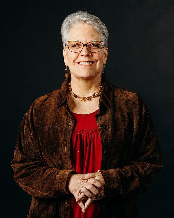 Woman in glasses, smiling, wearing a red top and brown jacket, hands clasped, against a dark background.