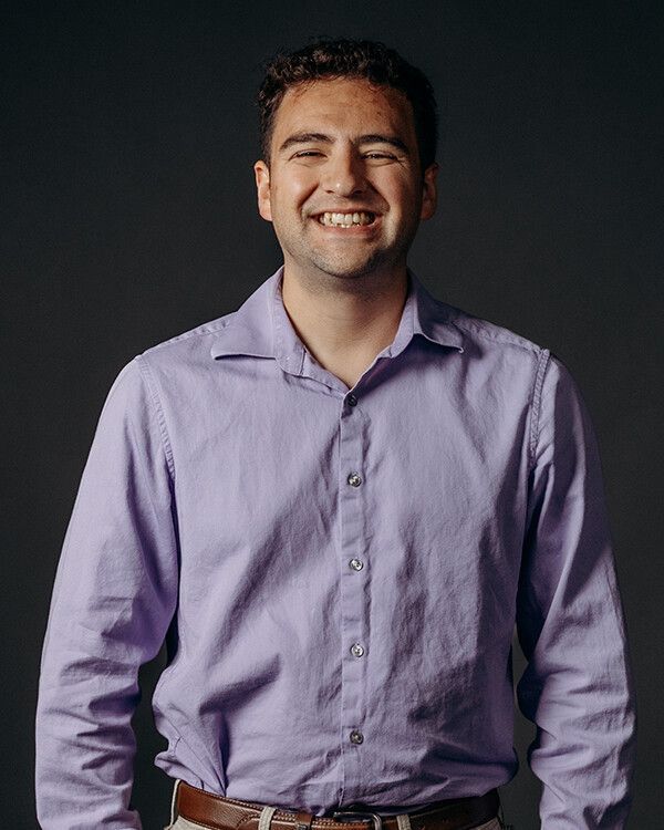 Man wearing a purple button-down shirt smiles in front of a gray background.