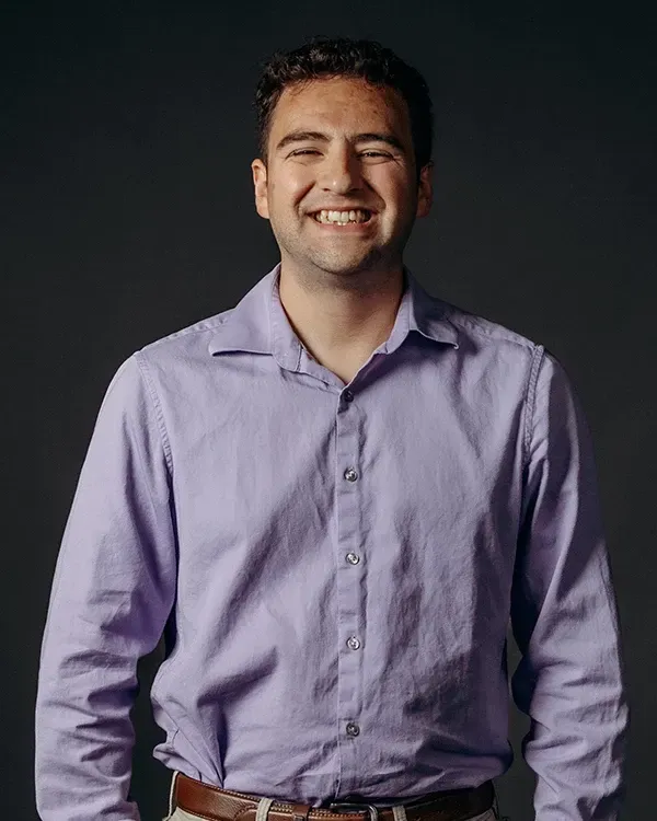 Man in purple button-down shirt smiles at the camera against a gray backdrop.