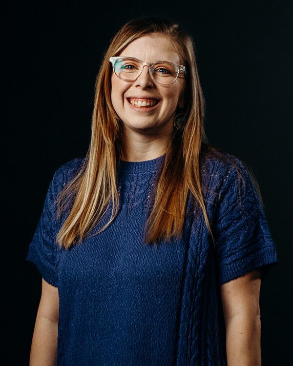 Woman with glasses smiling, wearing a blue sweater, against a black background.