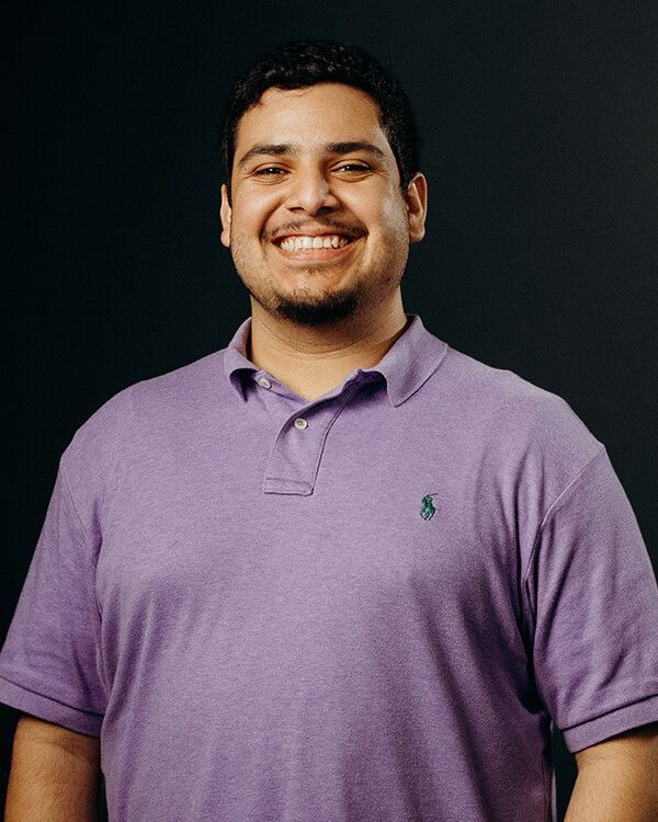 Man smiling, wearing a purple polo shirt, against a dark background.