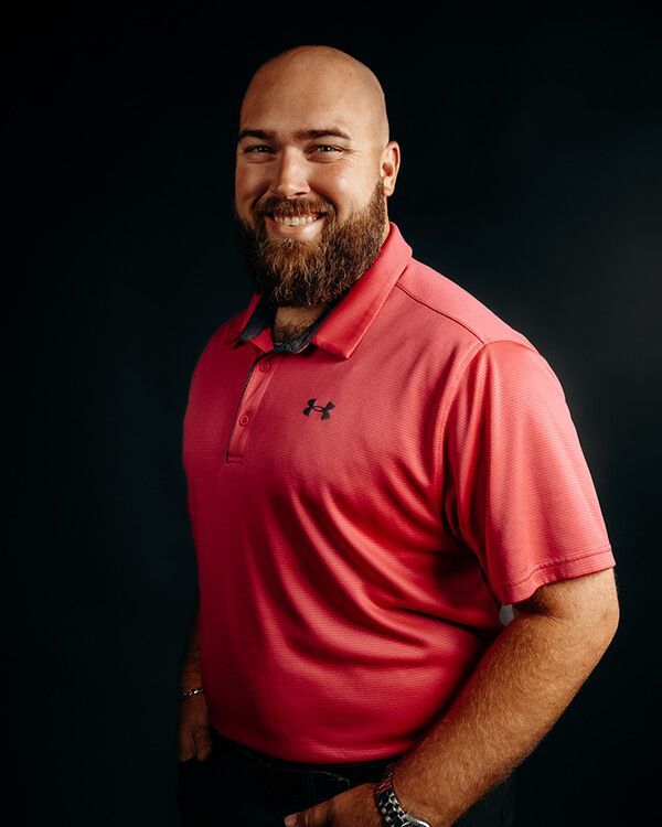 Man with a beard smiling, wearing a red polo shirt, against a black background.