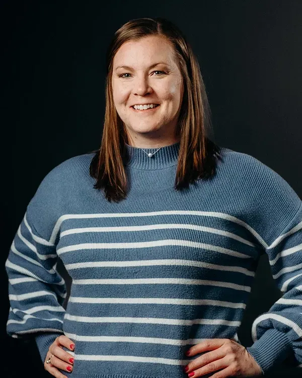 Woman smiling, wearing a blue and white striped sweater, hands on hips, dark background.