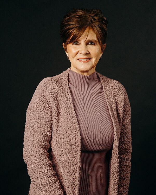 Woman in mauve sweater and cardigan smiles in front of a dark backdrop.