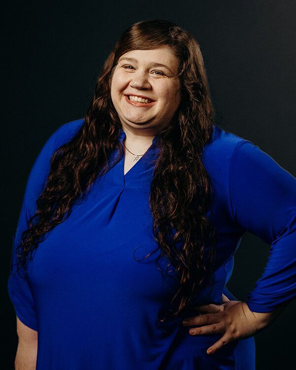 Woman in a blue top smiling with dark curly hair. Hand on hip, against a dark backdrop.
