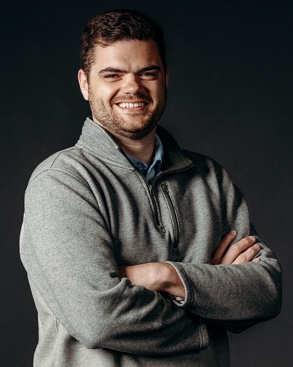 Man with crossed arms, smiling, wearing gray sweater over a blue collared shirt. Dark gray backdrop.