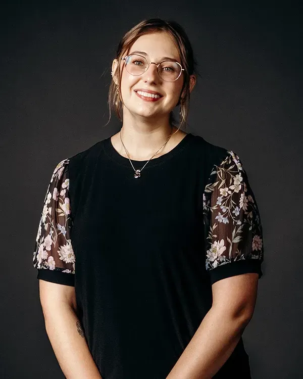 Woman with glasses smiles; wearing black top with floral sleeves. Gray backdrop.