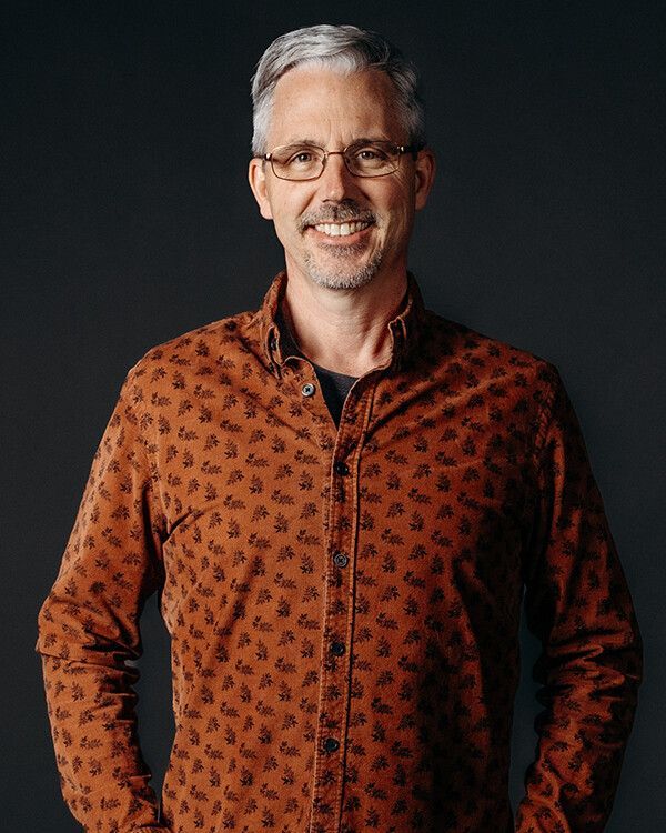Man wearing glasses and patterned shirt smiles, hands in pockets. Dark background.