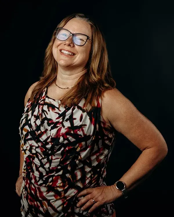 Woman with glasses smiling, wearing a sleeveless patterned top, posing with hands on hips, black background.