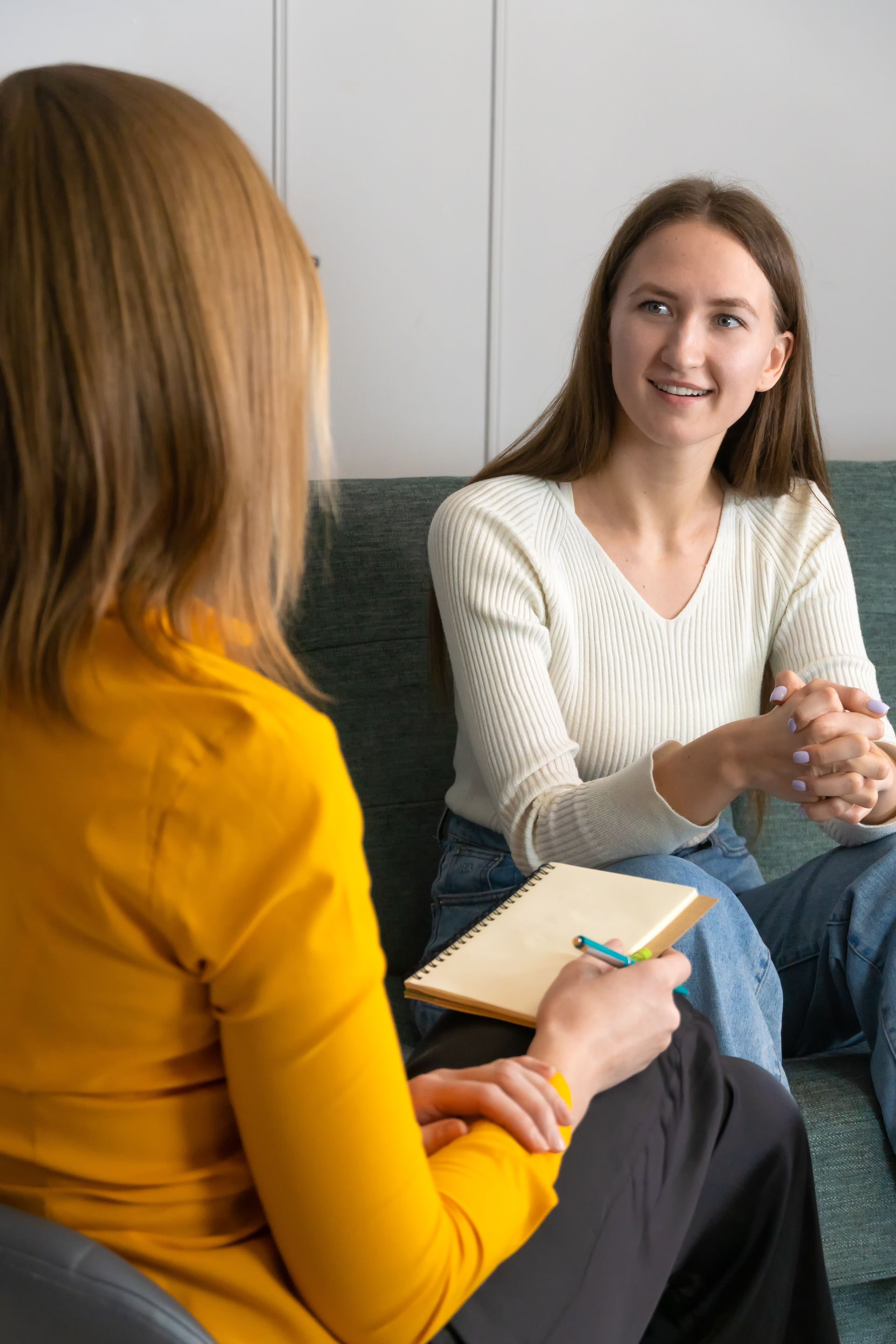 Woman in yellow shirt and other woman in white shirt having a conversation on a couch, one holding a notepad.