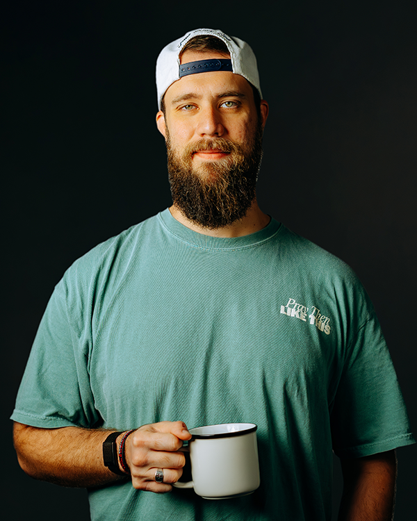 Man with beard smiles in a blue button-down shirt against a dark background.