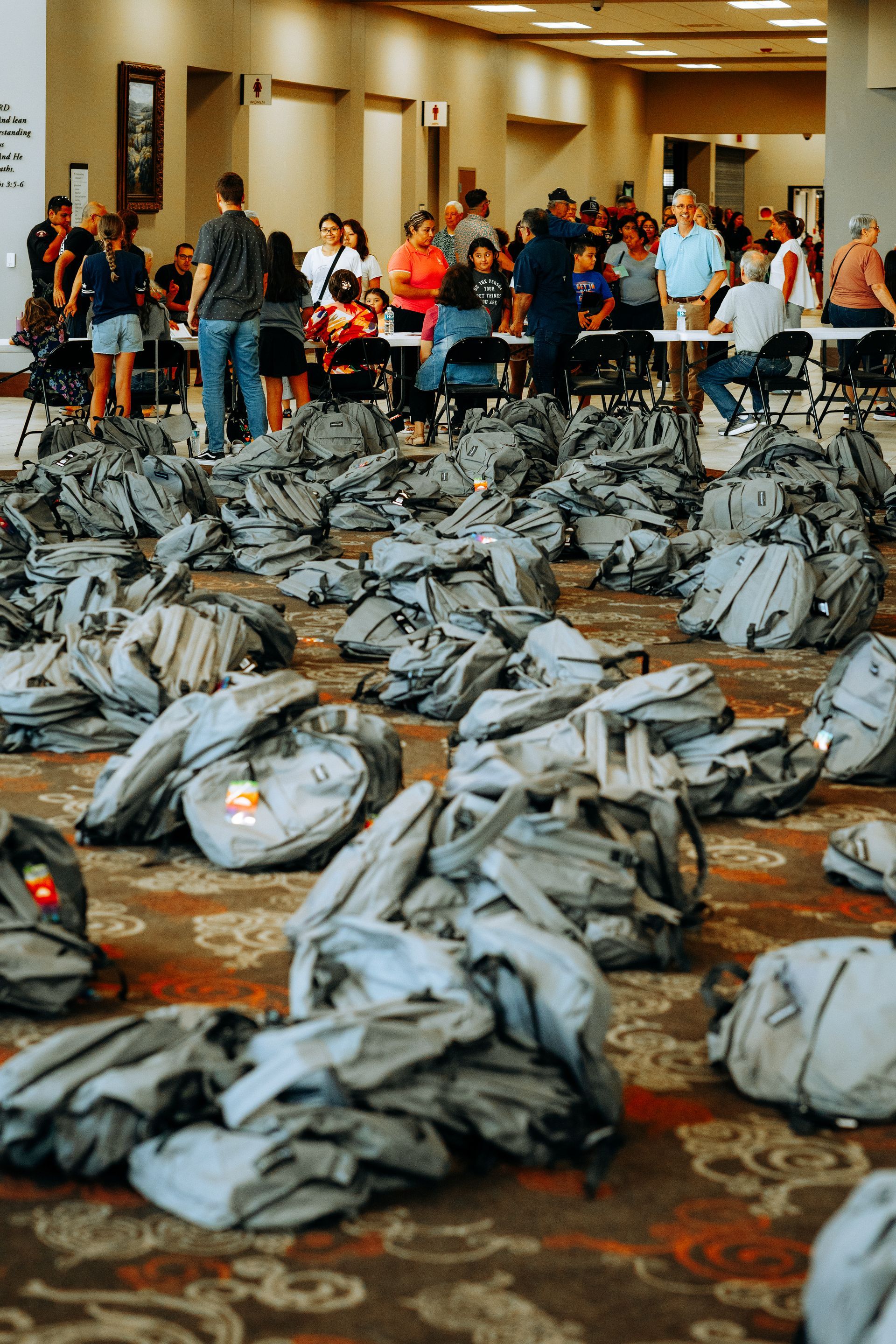 Floor covered in backpacks, people in background at tables. Large room, presumably indoors.