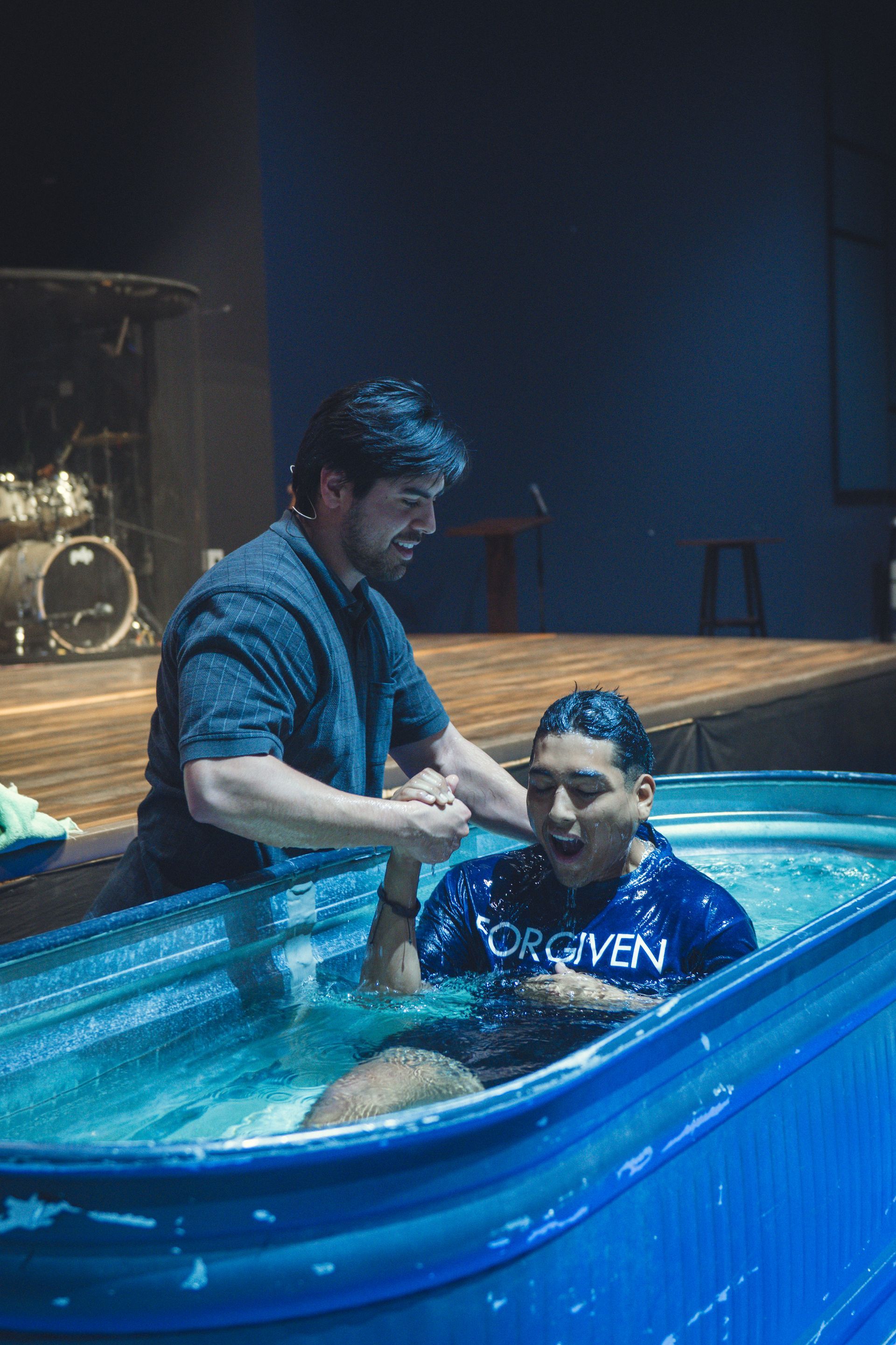 Person being baptized in a pool by another person; indoor setting with a drum set in the background.