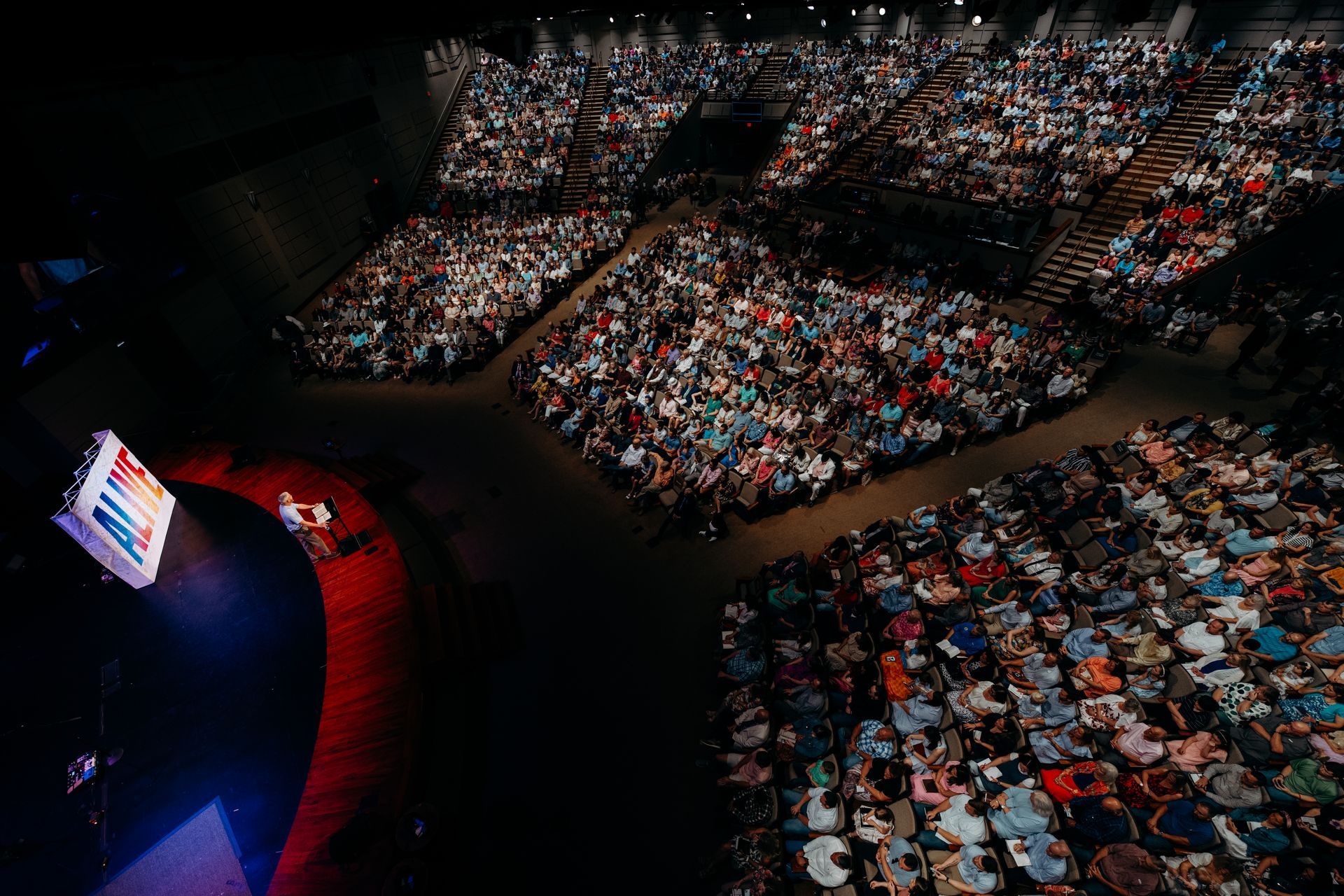Audience in large venue watching presentation on stage with screen.