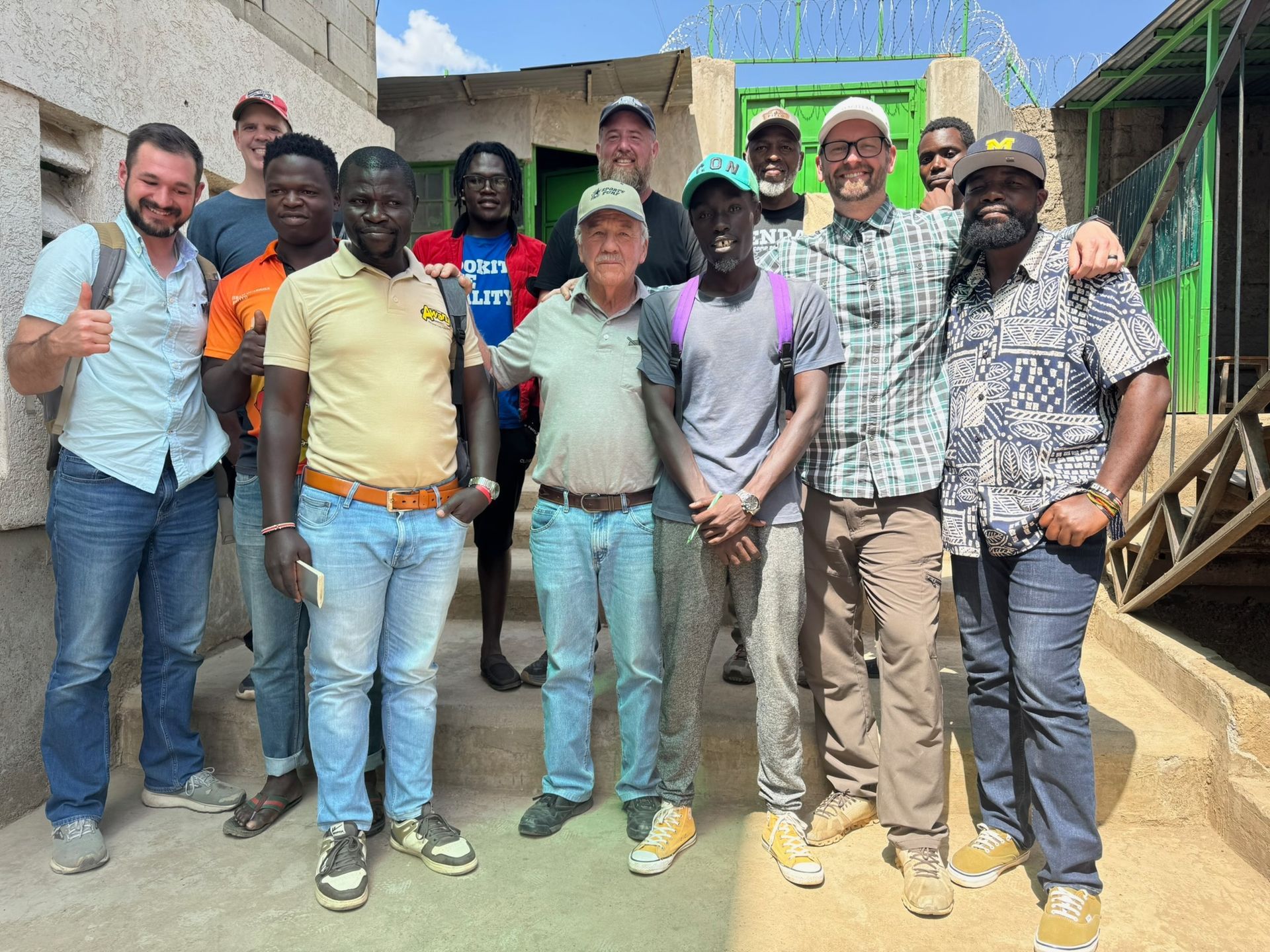 Group of people posing outdoors, smiling. Several wear caps; some have arms around each other. Buildings in the background.