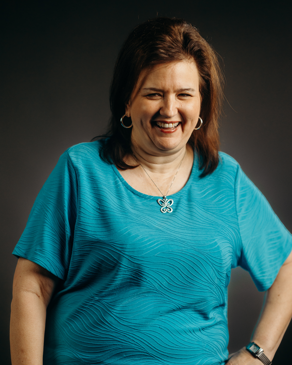 Woman in a turquoise top smiles, standing in front of a gray backdrop, arm on hip.