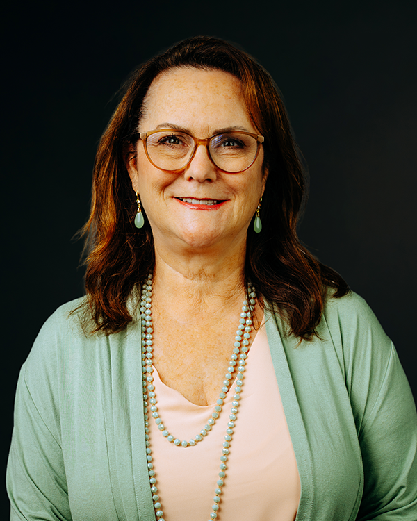 Woman with glasses and long hair smiles, wearing a black top and silver necklace against a dark background.