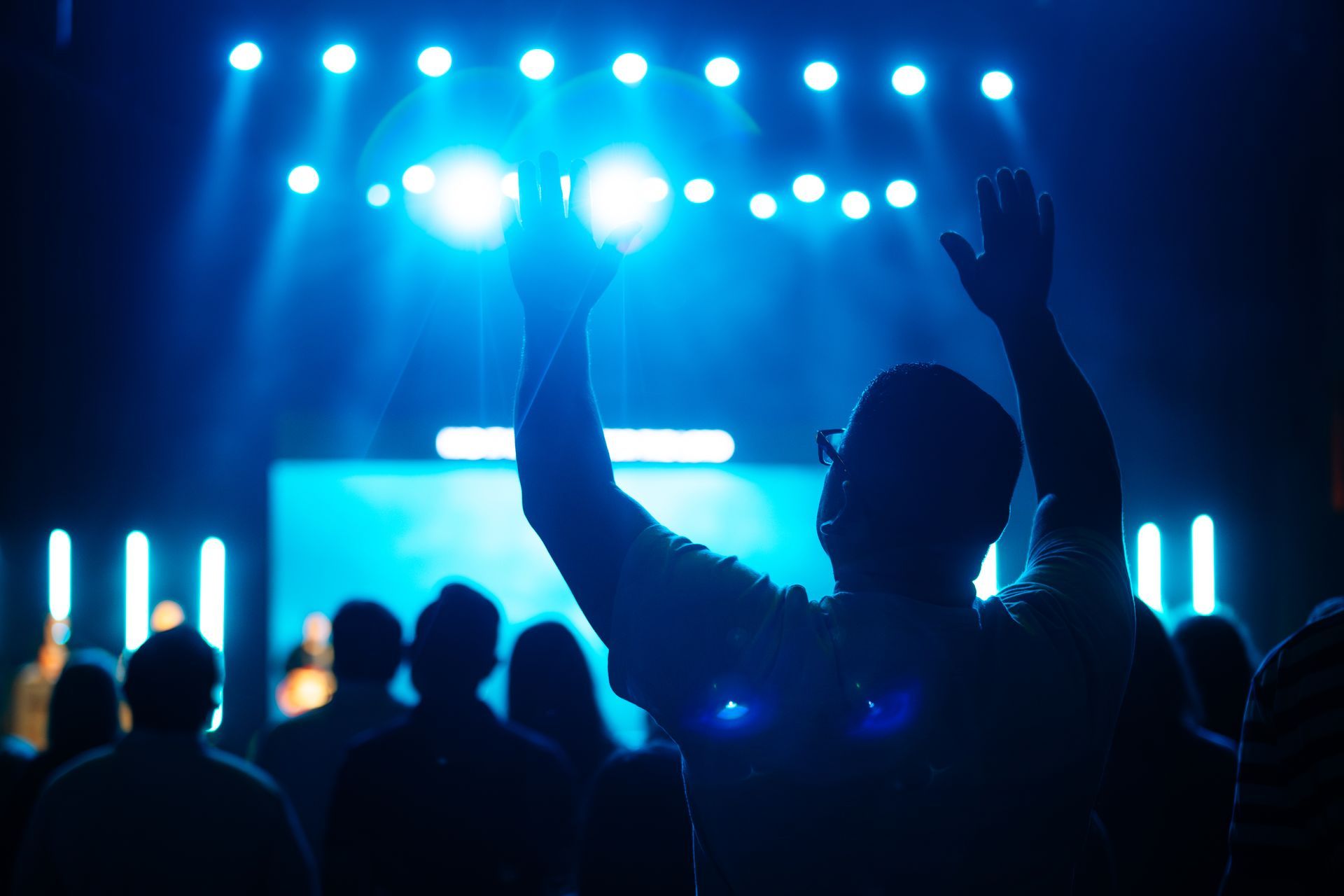Person with arms raised in worship at a concert with bright blue stage lights and a crowd.