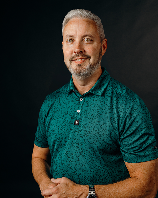 Man with graying hair and beard in a teal polo shirt smiles, dark backdrop.