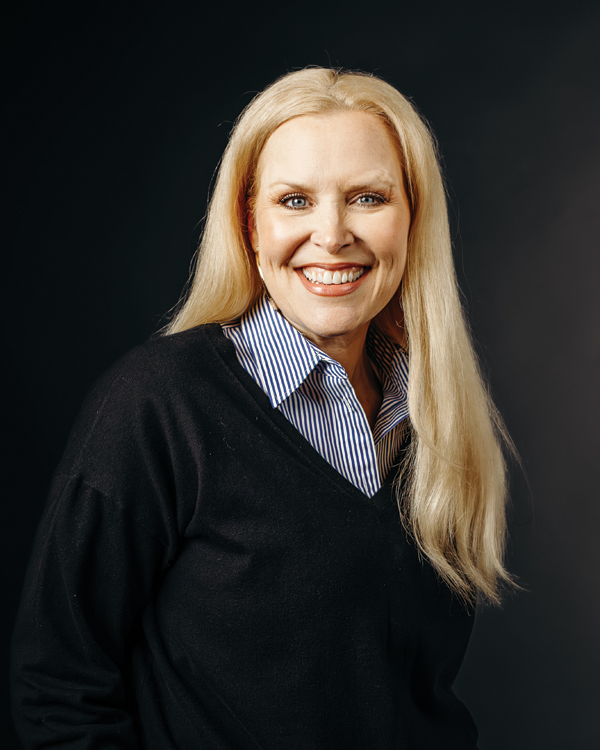 Woman with long blonde hair smiles, wearing a striped shirt under a black sweater. Black background.