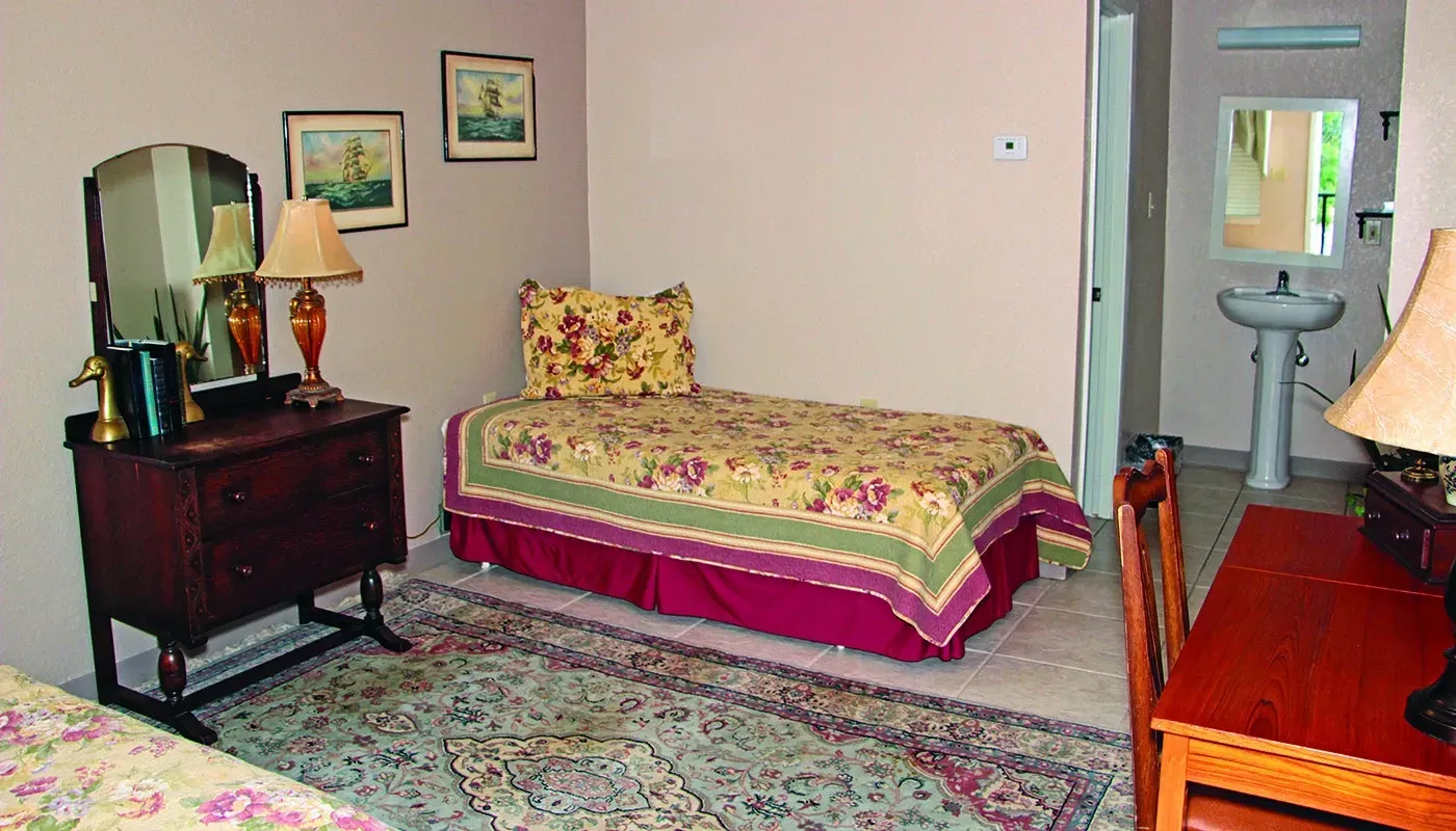 Bedroom with a bed, dresser, and a sink. The bed has a floral comforter and a burgundy skirt.
