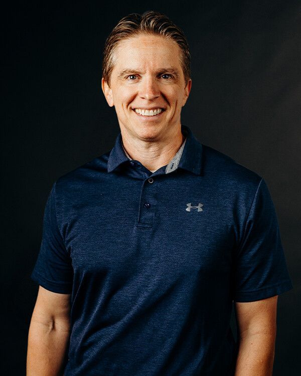 Man smiling, wearing a blue Under Armour polo shirt, against a dark background.