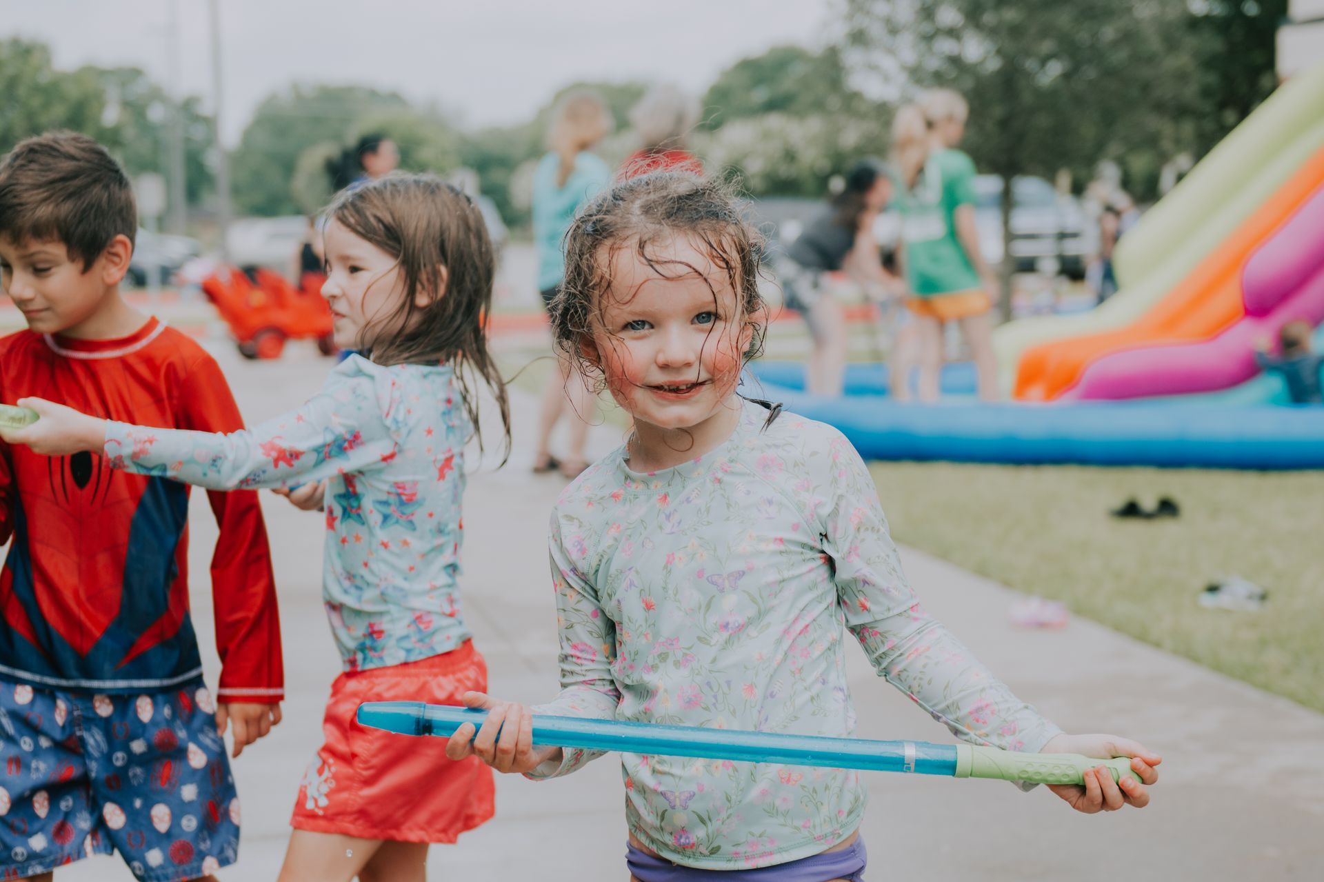 Children playing with water guns near a pool and inflatable slide. One child smiles, holding a water gun.