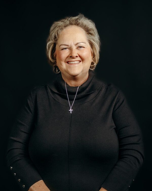 Woman smiling, wearing a black turtleneck and cross necklace, against a black background.
