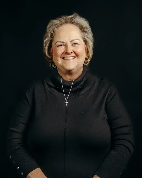 Woman with blonde hair, wearing a black turtleneck and cross necklace, smiles against a black background.