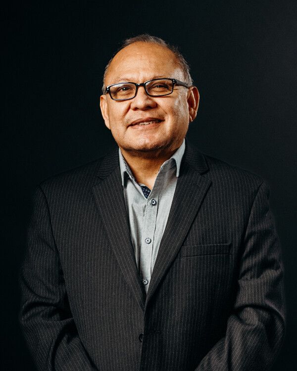 Man in glasses and suit jacket, standing against a dark background, smiling.