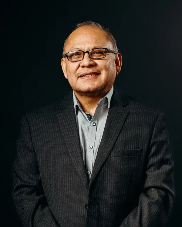 Man wearing glasses and a suit, smiling in front of a dark background.