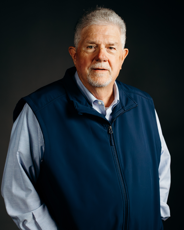 Man in navy vest and blue shirt, hands in pockets, looking forward against a gray backdrop.