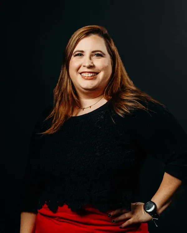 Woman in black top and red skirt, smiling, hand on hip, wearing a watch. Dark background.