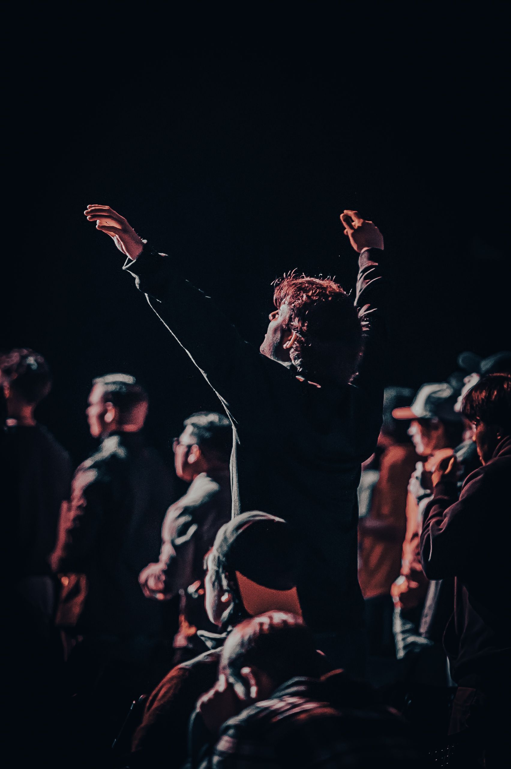 Person with arms raised in a crowd, illuminated by stage lighting. Dark setting, blurred figures.