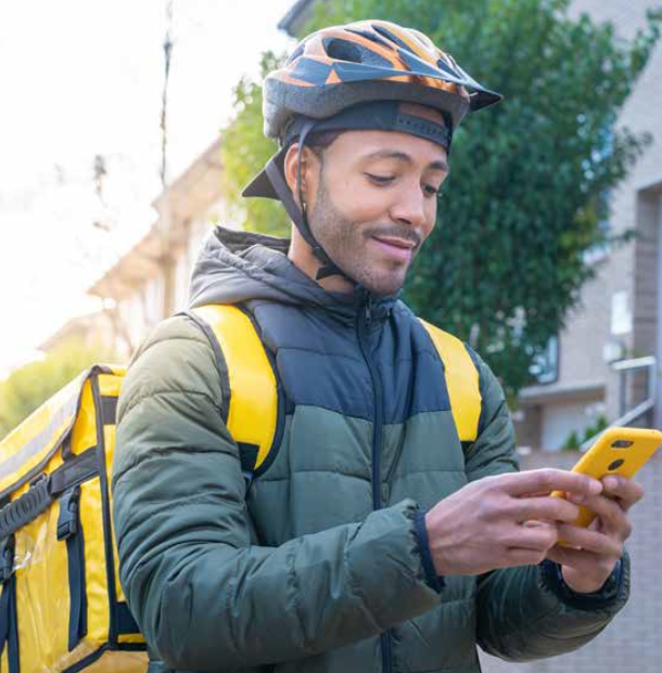 Delivery person on bicycle, wearing helmet and yellow backpack, looking at phone.