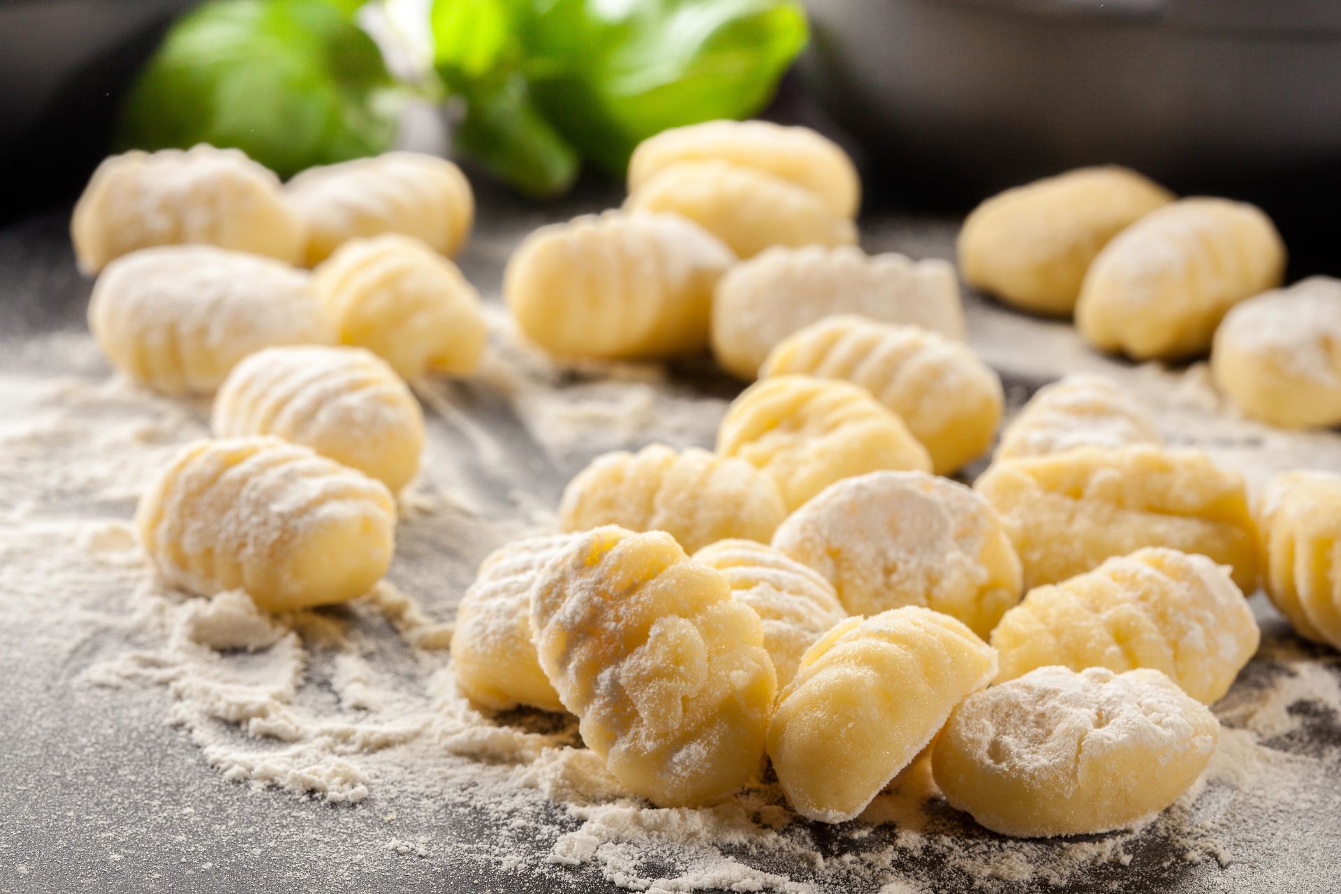 Gnocchi freschi crudi, infarinati, su un piano di lavoro scuro con erbe aromatiche sullo sfondo.