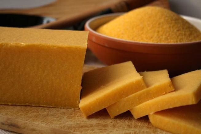 Fette di polenta gialla su un tagliere di legno con una ciotola di farina di mais gialla a grana grossa sullo sfondo.