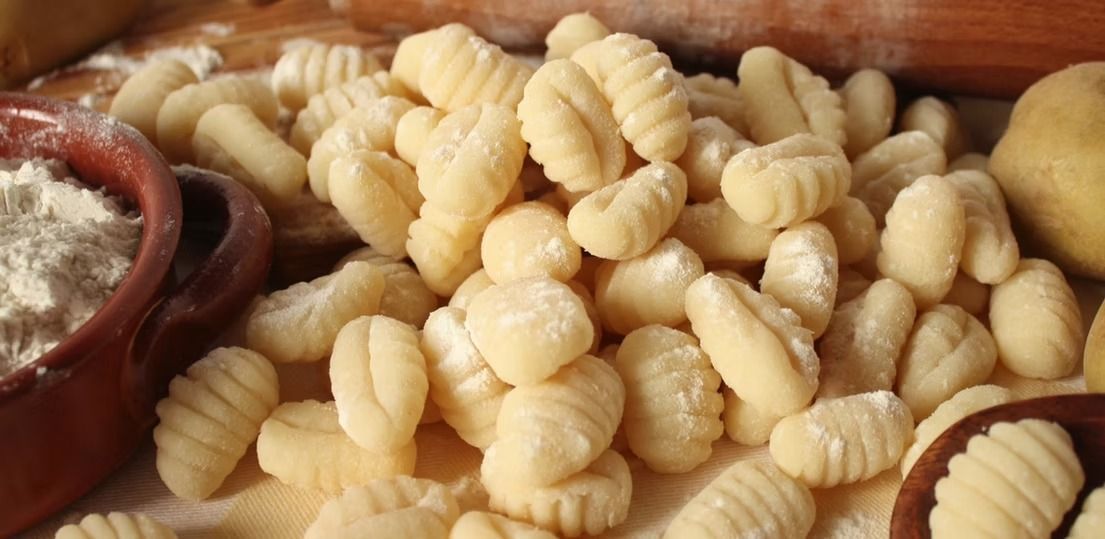 Un mucchio di gnocchi di patate crudi su un tagliere di legno, con una ciotolina di salsa nelle vicinanze.