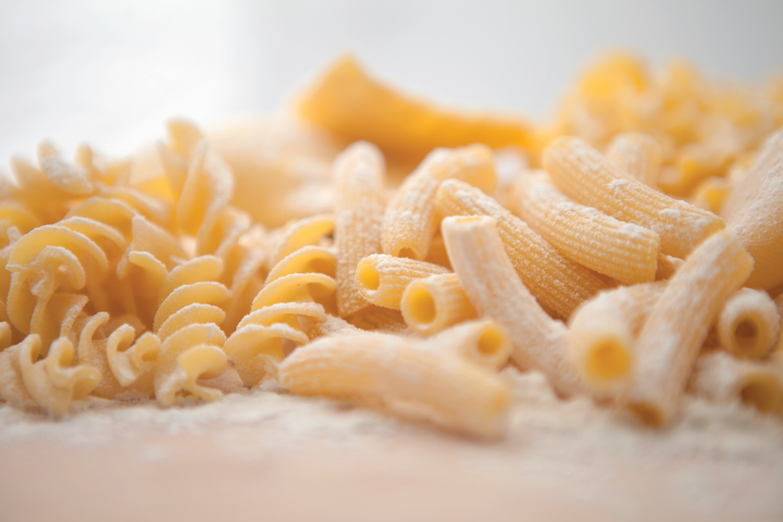 Vari formati di pasta cruda, tra cui fusilli, penne e conchiglie, disposti su una superficie bianca in una luce calda.
